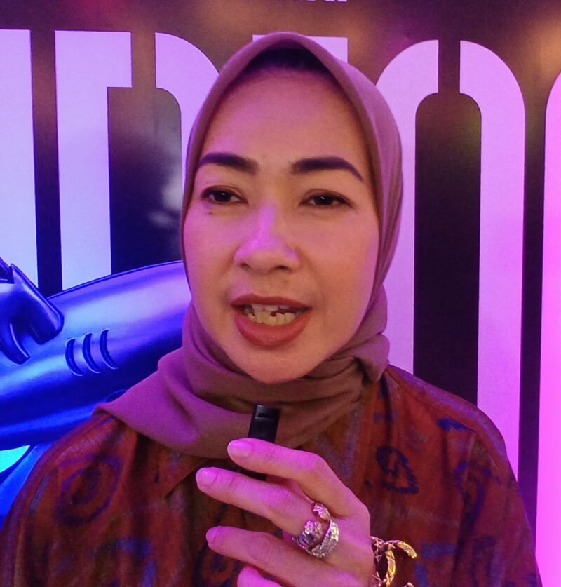 Foto: Alia Rohali terlihat berdiri di depan latar bertema Flying Fish Alumni sambil memegang mikrofon kecil.