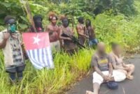 Foto: Sejumlah pria bersenjata berdiri di tepi Jalan Trans Papua, Distrik Seradala, Kabupaten Yahukimo, Papua Pegunungan, sementara dua warga sipil dengan wajah disamarkan terlihat duduk di depan mereka. 
