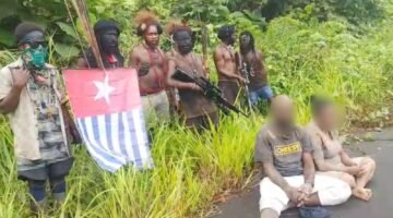 Foto: Sejumlah pria bersenjata berdiri di tepi Jalan Trans Papua, Distrik Seradala, Kabupaten Yahukimo, Papua Pegunungan, sementara dua warga sipil dengan wajah disamarkan terlihat duduk di depan mereka.