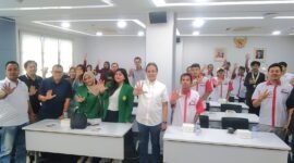Foto: Peserta kegiatan bersama narasumber dan tamu undangan berfoto bersama dengan mengangkat tangan di ruang pertemuan.