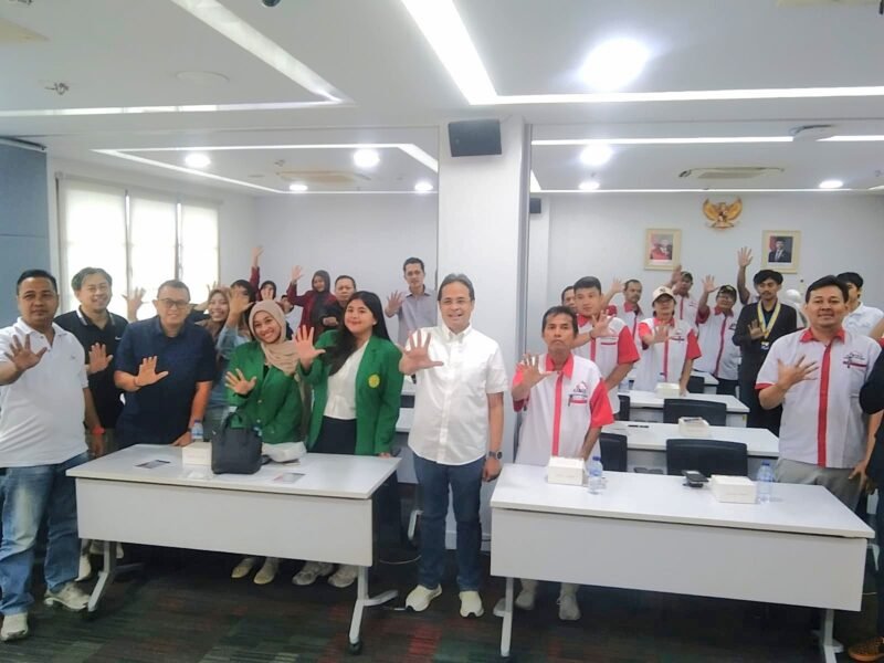 Foto: Peserta kegiatan bersama narasumber dan tamu undangan berfoto bersama dengan mengangkat tangan di ruang pertemuan.
